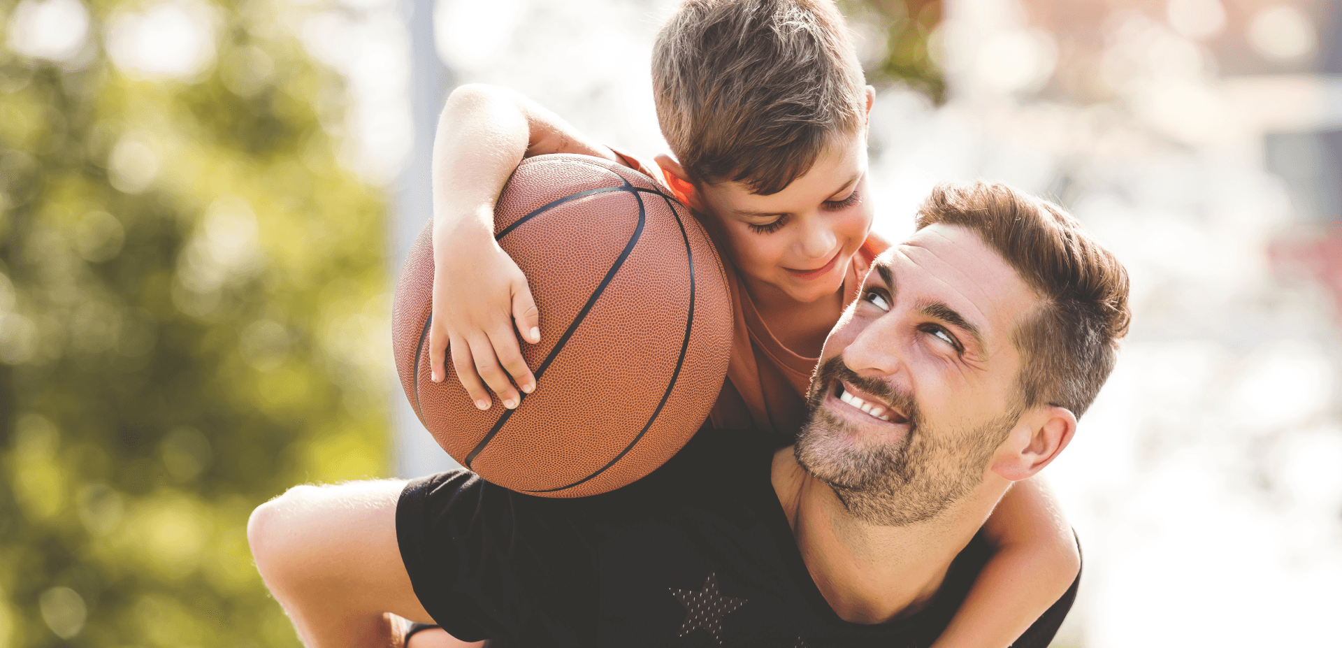 dad looking up at young boy riding piggyback holding basketball and smiling at each other