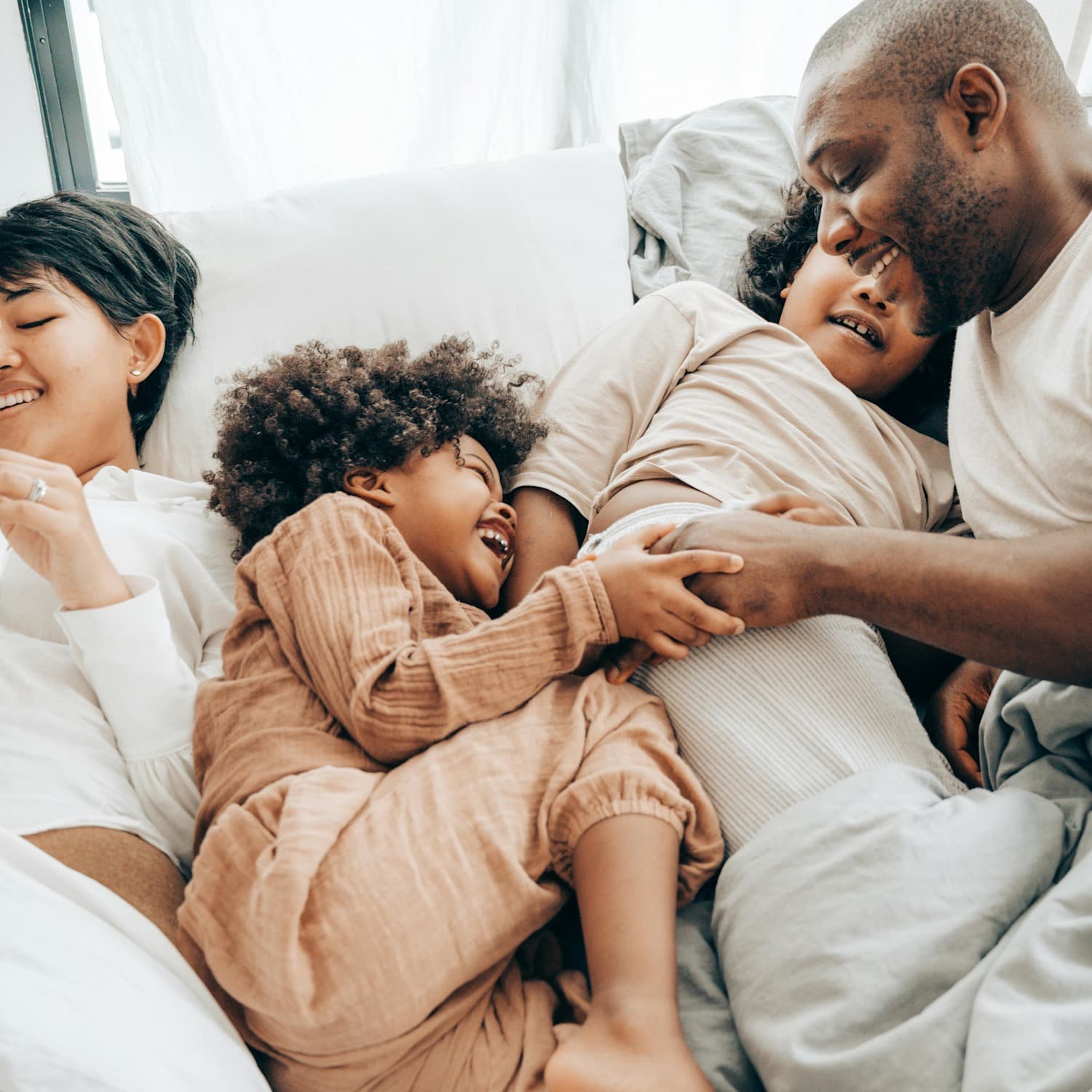 A father, two young kids, and a mother are all smiling and snuggling in bed, cozy with blankets