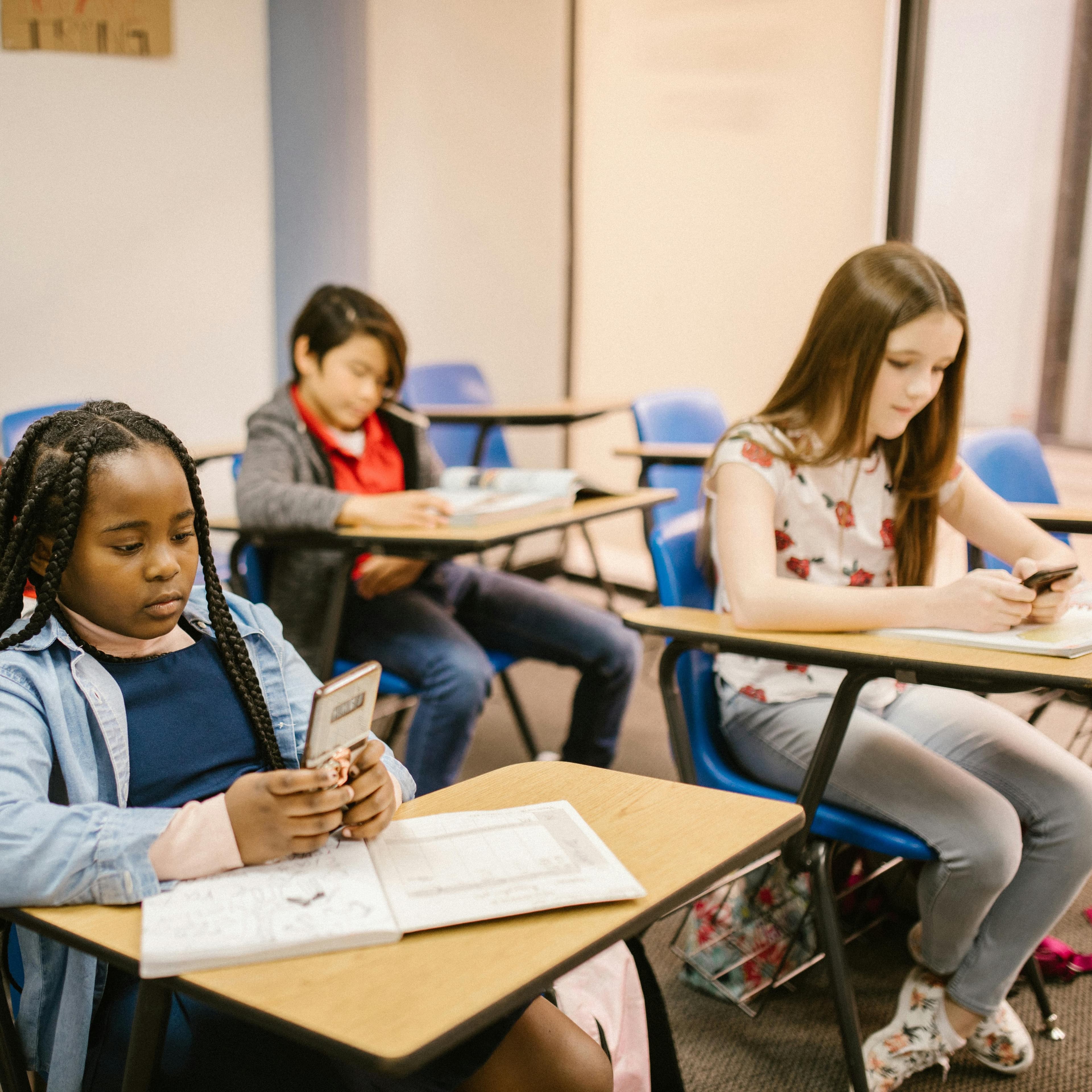 Three elementary age kids sitting at school desks looking at their phones