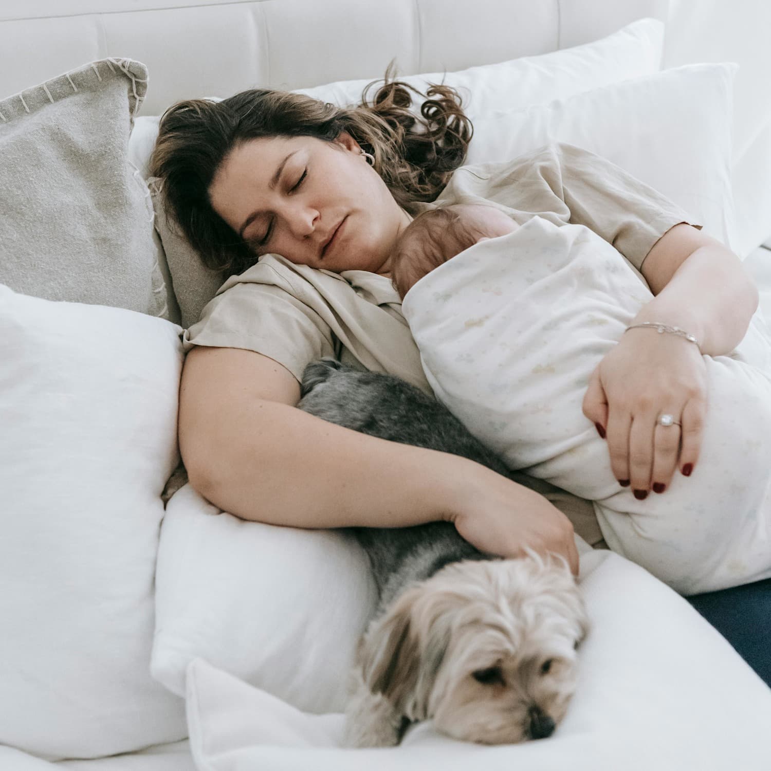 A mother falls asleep on the couch holding a sleeping baby wrapped in a blanket and petting a dog