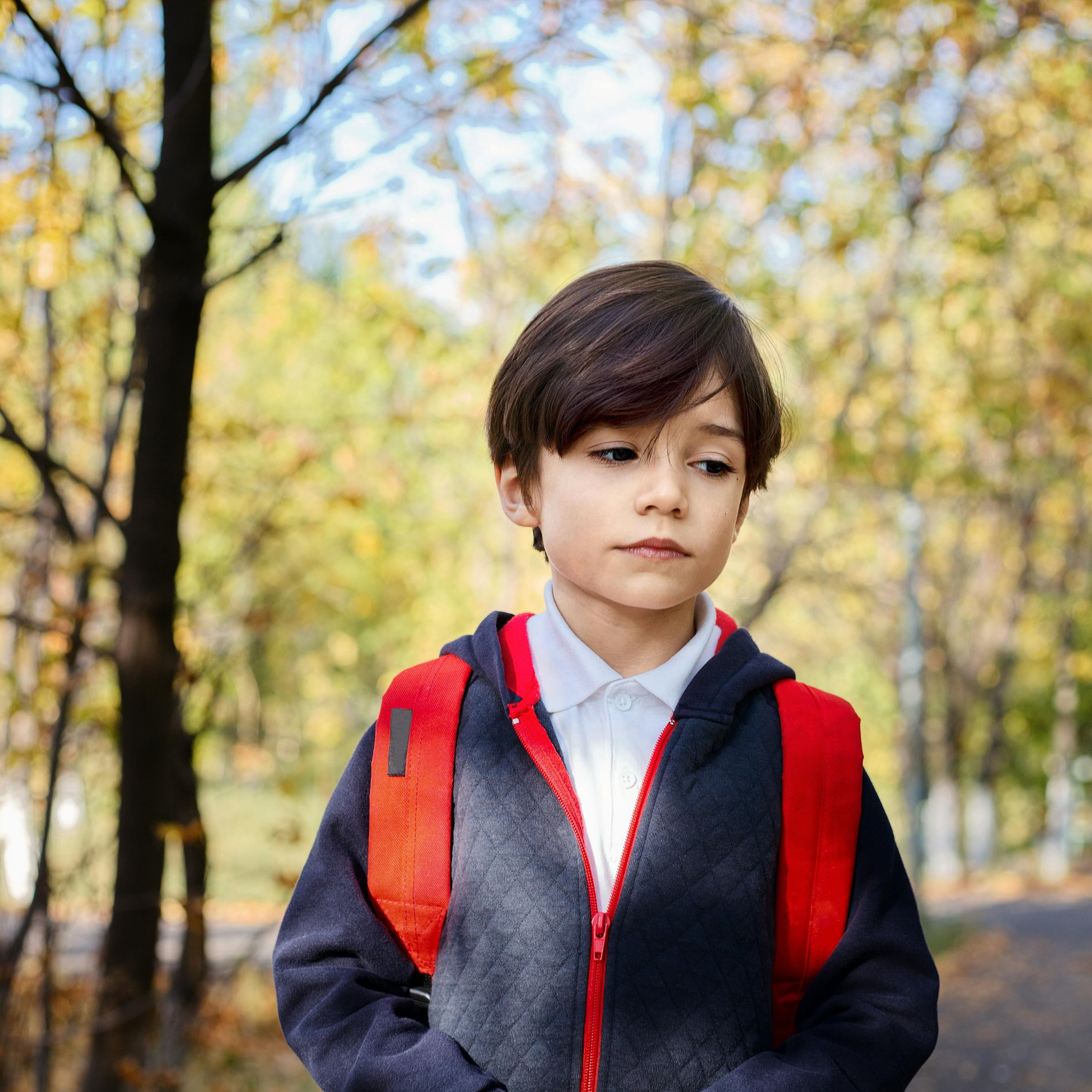School-aged boy walking in woods with backpack and jacket on and a forlorn look