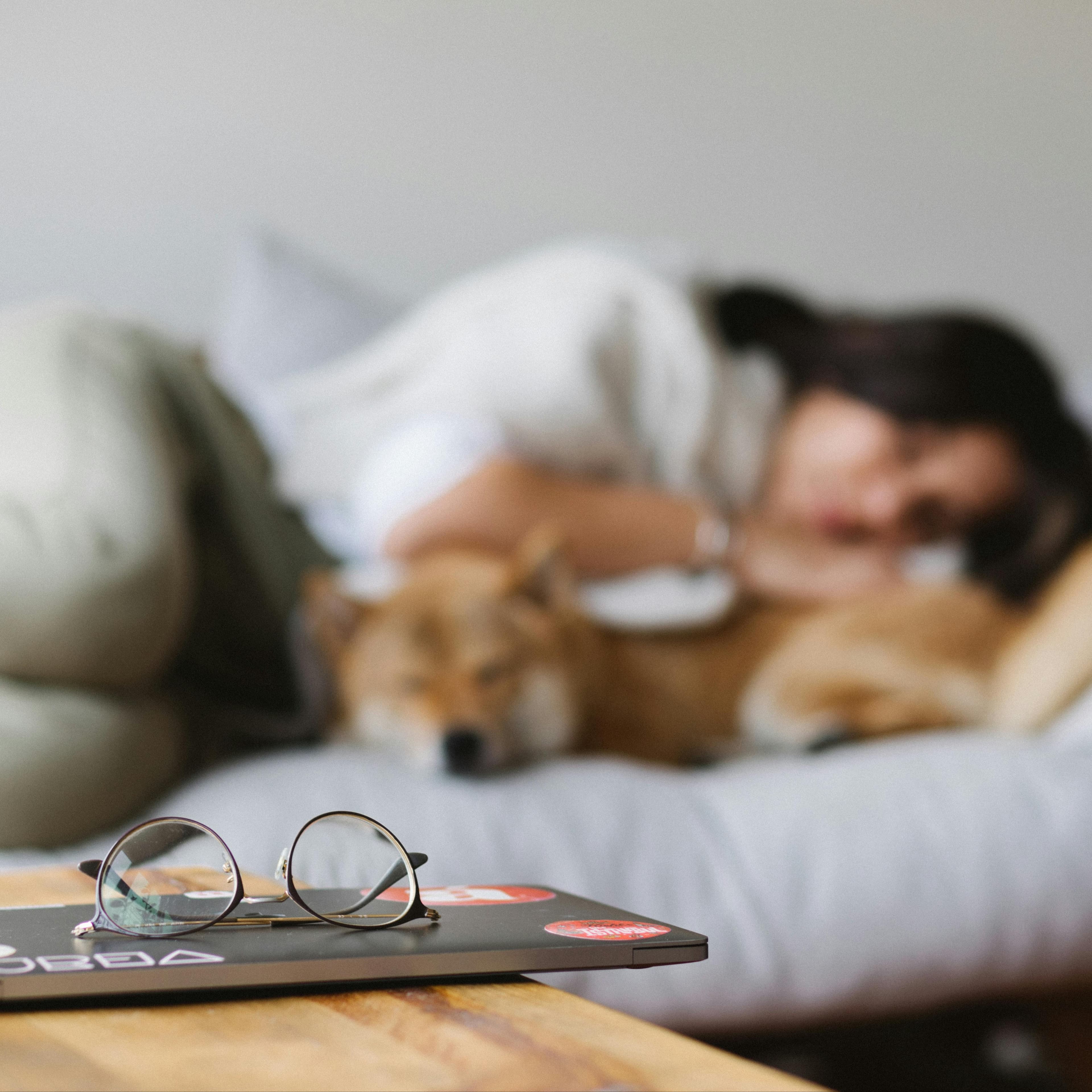 Young woman curled up on bed with dog, blurry in the background, closed computer and glasses on desk in foreground