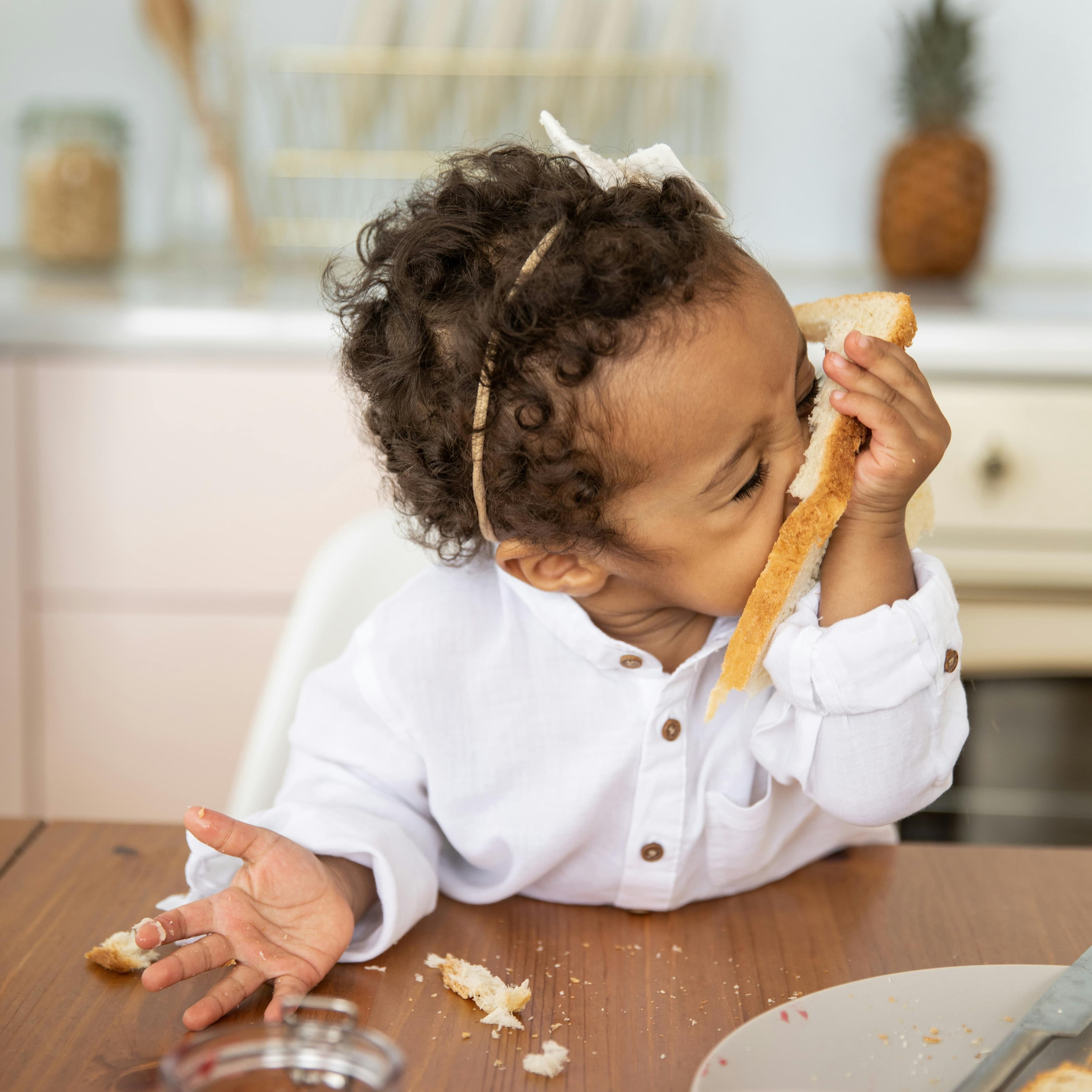 Toddler girl with a white sweater and headband sitting at a table with crumbs eating a piece of toast and making a mess