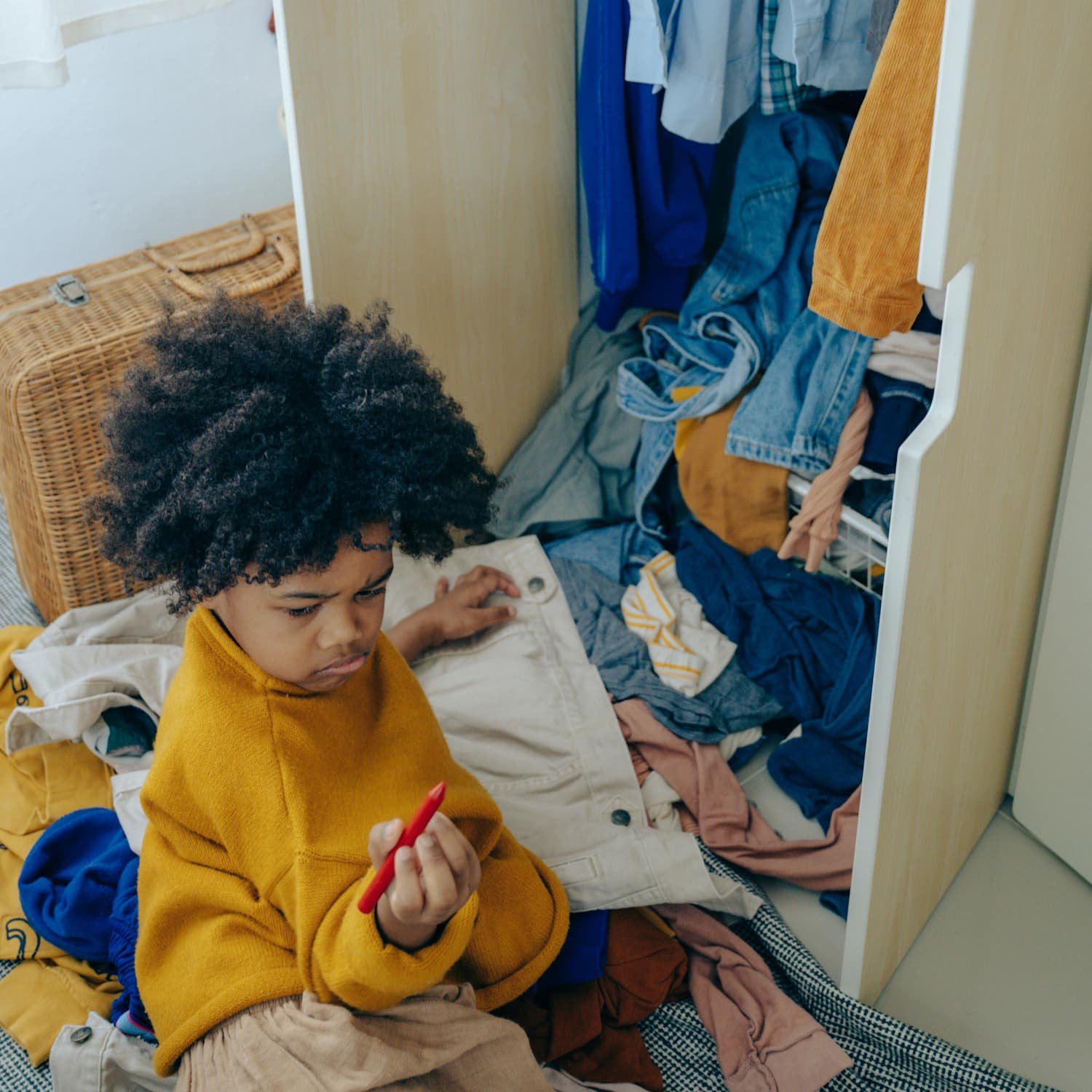 Young child sitting at the foot of a messy closet with clothes all around them, holding and looking at a red crayon